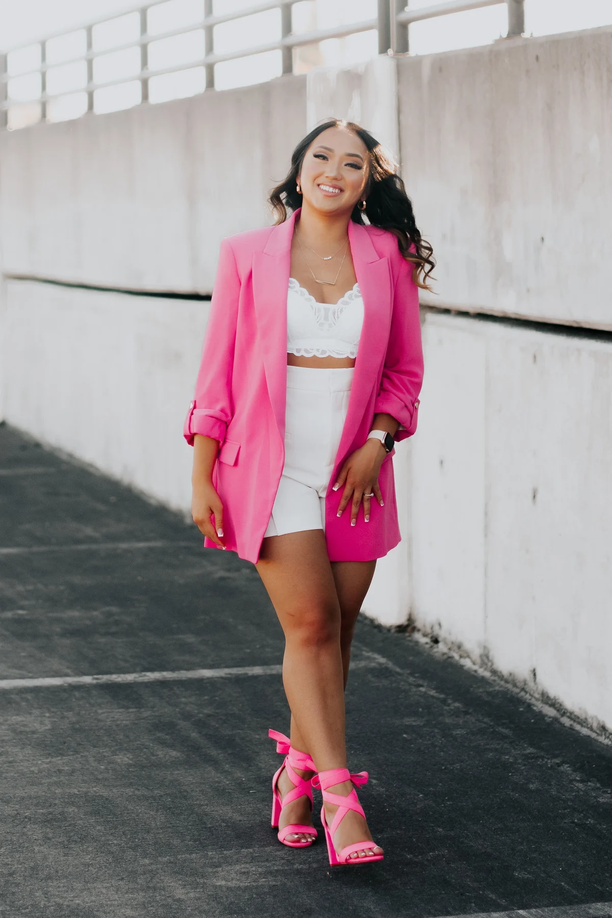 Rikki in a pink blazer, white top and shorts and pink lace-up heels, walking along a rooftop deck